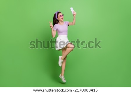 Full size funny photo of cool brunette lady jump do selfie wear head band glasses t-shirt skirt sneakers isolated on green background