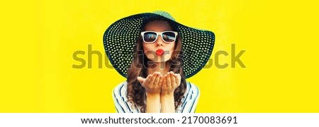 Portrait of beautiful young woman blowing her lips sending air kiss wearing sunglasses, summer straw hat on yellow background