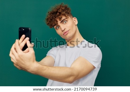 a joyful, handsome man takes a selfie on his smartphone holding it with both hands and smiling broadly. Horizontal studio photo on a green background