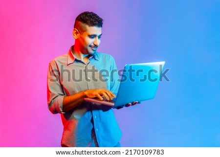 Portrait of man in shirt holding laptop in hand and typing, blogger making posts in social networks, chatting with followers. Indoor studio shot isolated on colorful neon light background.