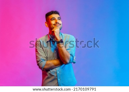 Portrait of man in shirt standing, touching his chin, toothy smile and looking away dreaming, imagining and fantasizing. Indoor studio shot isolated on colorful neon light background.