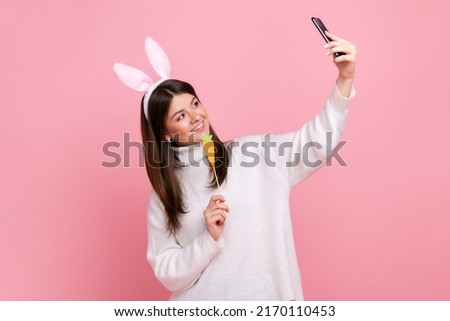 Side view portrait of funny girl with bunny ears streaming or has video call with carrot in hands, wearing white casual style sweater. Indoor studio shot isolated on pink background.
