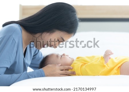 asian mom seeing and playing with her baby child who is sleep on bed, mom are smiley look happy in the morning in conceptual mother's day.