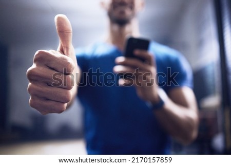 This personal trainer approves this app. Shot of an unrecognisable man showing using a smartphone and showing thumbs up in a gym. Royalty-Free Stock Photo #2170157859