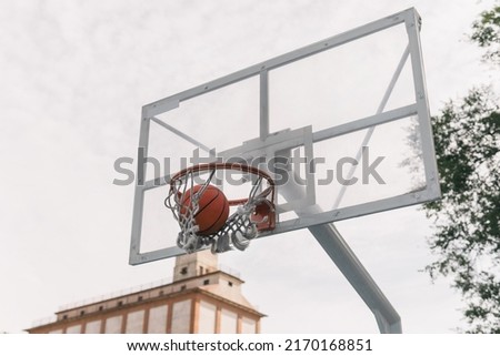 Basketball basket with an orange hoop and broken white nets with a ball going in. Glass or transparent plastic sports board to play basketball seen from below. Side and low angle view. Sport concept