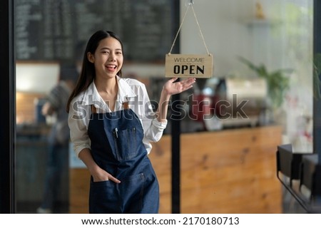 Happy waitress stands at the entrance of the restaurant showing off a sign of the store open. persuade customers to enter the store The smiling owner in a blue apron stood at the entrance to the cafe