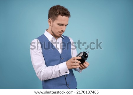 Young business man stands with an alarm clock in his hands on a blue studio background. The concept of using the right time