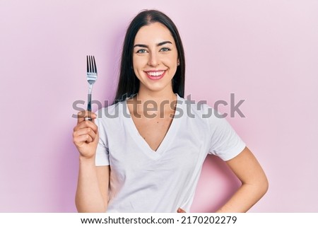 Beautiful woman with blue eyes holding one silver fork looking positive and happy standing and smiling with a confident smile showing teeth 