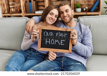 Young caucasian couple holding blackboard with our first home message sitting on the sofa.