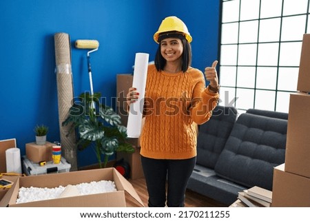 Young hispanic woman standing on new home with blueprints smiling happy and positive, thumb up doing excellent and approval sign 