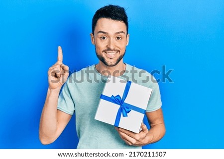 Young hispanic man holding gift smiling with an idea or question pointing finger with happy face, number one 