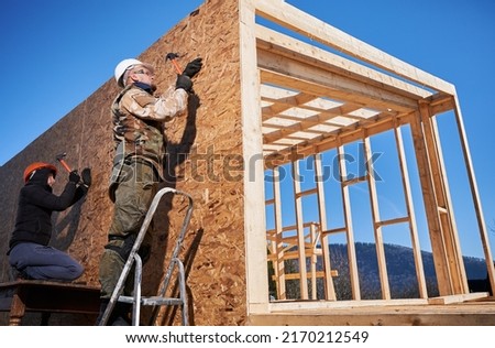 Carpenters hammering nail into OSB panel on the wall of future cottage. Men workers building wooden frame house in the Scandinavian style barnhouse. Carpentry and construction concept.