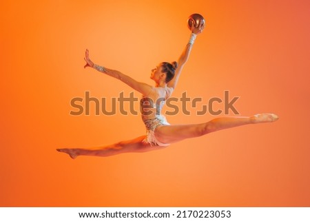 Leap, jump. One professional rhythmic gymnastics artist training with golden color ball isolated on orange background. Concept of sport, action, ad, aspiration, education, active lifestyle