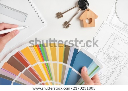 Hands choosing Colors of Interior Design from the Color Guide. Apartment Sketch, Paint palette, Model of a House and Keyring on the Background. Architect Using Color Swatch. House Plan with Blueprints