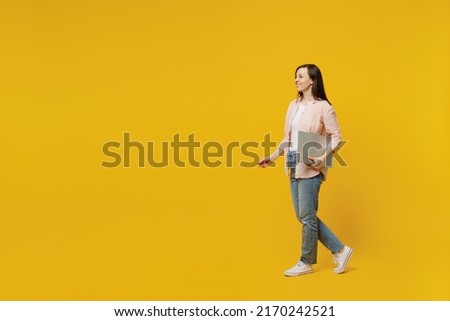 Full body side view young happy smiling woman she 30s wears striped shirt white t-shirt hold use closed laptop pc computer walk go isolated on plain yellow background studio. People lifestyle concept