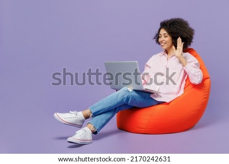 Full body young woman of African American ethnicity 20s wear pink striped shirt sit in bag chair hold use work on laptop pc computer waving hand isolated on plain pastel light purple background studio