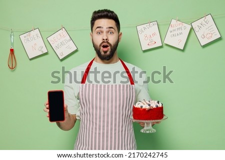 Young shocked male chef confectioner baker man 20s in apron hold birthday sweet dessert cake mobile cell phone with blank screen workspace area isolated on plain green background Cooking food concept