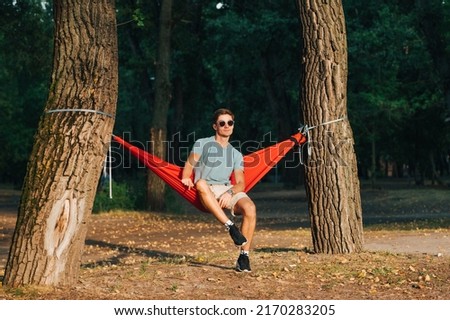 Handsome man in bright stylish clothes sits in a hammock in nature in the evening at sunset on a background of forest and looks away with a serious face. Tourist on a hammock resting in the woods. Royalty-Free Stock Photo #2170283205