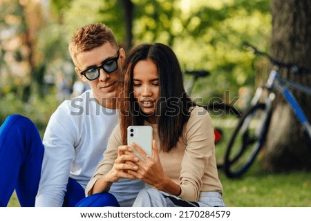 Pretty hispanic female is holding a smartohone, texting, showing screen to her boyfriend. The couple is watching on the phone, sitting in the park. Royalty-Free Stock Photo #2170284579