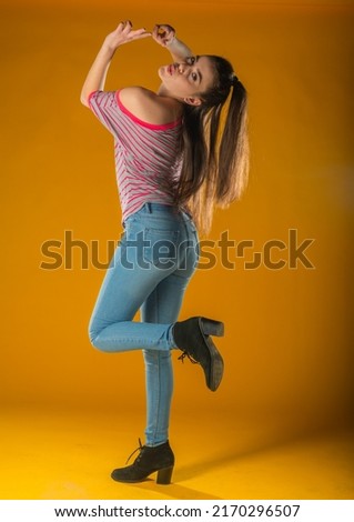 Beautiful young woman in pink mini dress posing with hand on hip, presenting something and looking away. Three quarter length studio shot on yellow background.