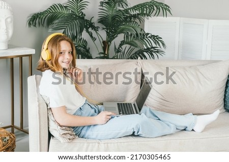 Teenage girl in yellow headphones smiles. Young redhead beauty. A red-haired girl uses a laptop and headphones to study. A teenager is resting at home on the couch. Home schooling
