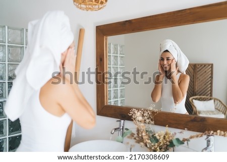 Young beautiful female with towel on head smiling to mirror reflection  standing in the bathroom at home. 30 years old happy woman doing daily morning rituals and cleansing. Enjoying healthy skin care Royalty-Free Stock Photo #2170308609