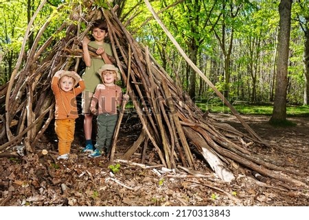 Three children boys and a girl with pile of brushwood in the forest build hut of branches in summer school Royalty-Free Stock Photo #2170313843