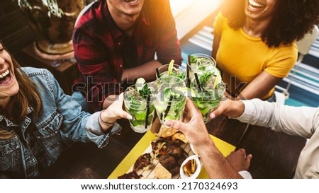 Multiracial friends cheering mojitos at bar restaurant - Young people celebrating happy hour toasting drinks and eating appetizers at tropical cocktail pub - Beverage and youth lifestyle concept