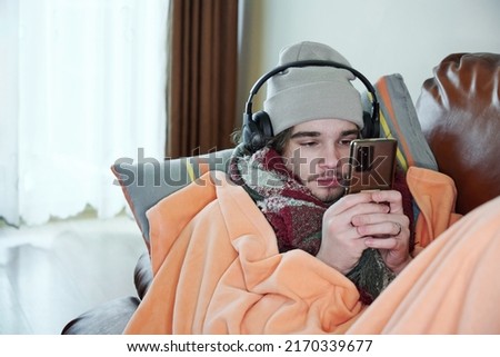 Young man warmly dressed is feeling cold, covered with warm blanket, sitting on the couch at cold home. Have problem with health, central heating Royalty-Free Stock Photo #2170339677