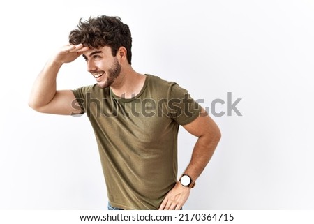Hispanic man standing over isolated white background very happy and smiling looking far away with hand over head. searching concept.  Royalty-Free Stock Photo #2170364715