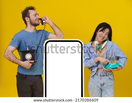 Handsome guy and asian girl talking on vintage phones leaned on huge smartphone or digital tablet with blank screen, happy smiling isolated on yellow background. Mock up, product placement. 