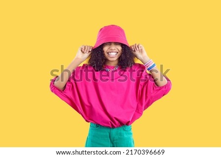 Happy joyful kid in trendy casual outfit puts pink bucket hat on her head. Cheerful beautiful African girl wearing loose fuchsia top and green pants having fun and laughing. Children's fashion concept Royalty-Free Stock Photo #2170396669