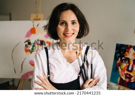 Real Female Artist Dirty with Paint, Wearing Apron, Crosses Arms while Holding Brushes, Looks at the Camera with a Smile. Authentic Creative Studio with Large Canvases. Head and Shoulders Portrait