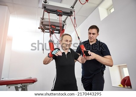 Rehabilitation specialist doing active treatment on suspension straps to elderly patient. Therapeutic exercises to restore pain-free movement patterns and improve function on red cord slings Royalty-Free Stock Photo #2170443295
