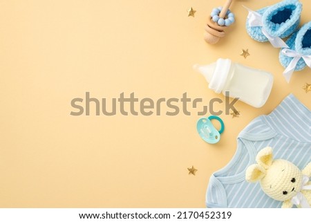 Baby accessories concept. Top view vertical photo of blue shirt knitted booties milk bottle wooden rattle soother knitted bunny toy and gold stars on isolated pastel beige background with empty space