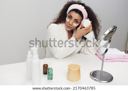 Flawless tanned curly Latin lady hold facial cream bottle looks at camera enjoying doing spa procedure in home interior. Copy space. Aesthetic medicine skincare beauty products ad concept