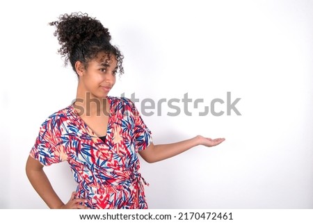 Portrait of young beautiful brunette woman wearing colourful dress over white wall  with arm out in a welcoming gesture.