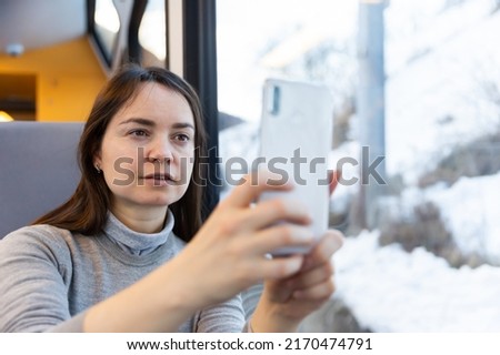 Dark-haired woman enjoying journey in modern comfortable train, looking with interest at picturesque scenery outside window and taking pictures on cellphone