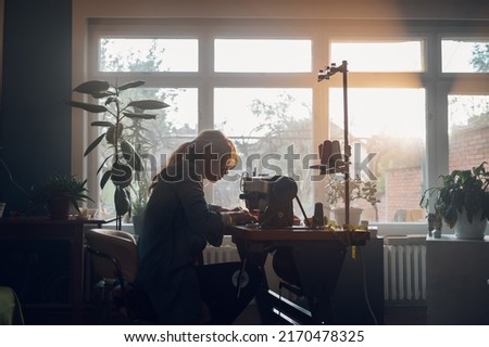 Silhouette of a beautiful middle aged tailor woman sewing on a sewing machine while sitting at her moody working place at home. Fashion atelier, tailoring, handmade clothes concept. Slow Fashion. Royalty-Free Stock Photo #2170478325