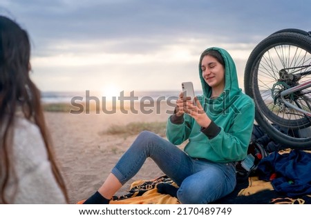 smiling latin young woman sitting on the beach outdoors taking pictures of her friend
