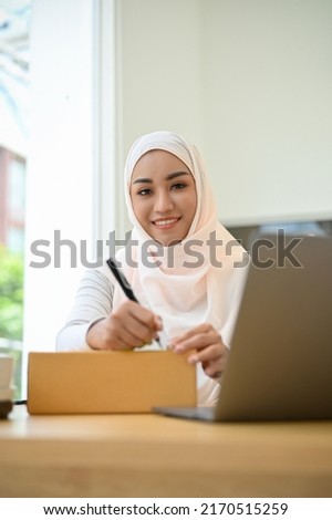 Charming asian muslim female small e-commerce business entrepreneur packing a shipping box, writing a customer address on a cardboard box.