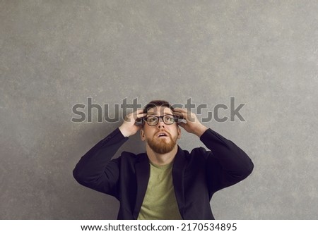 Young man afraid of something. Shocked scared anxious guy in hipster glasses looking up at terrible threat and danger sitting against grey concrete studio wall. Fear, fright, problem, trouble concept Royalty-Free Stock Photo #2170534895