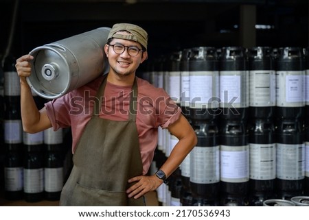 Male worker with metal beer kegs at a brewery Small Business and the Beer Industry Smiling handsome Asian man in an apron holding a steel bucket over his shoulder in a warehouse.
