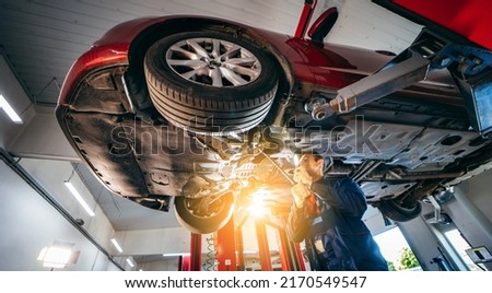 Young car mechanic at repair service station inspecting car wheel and suspension detail of lifted automobile. Bottom view. Royalty-Free Stock Photo #2170549547