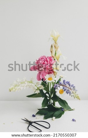 Modern summer flowers composition on rustic white table indoors. Creative floral image. Stylish peony, lupin,iris and daisy arrangement on kenzan with scissors on rustic wood.