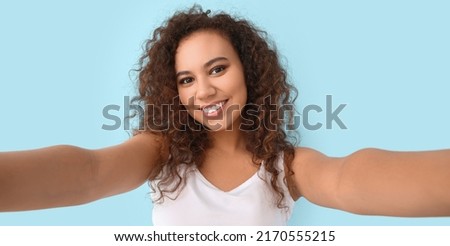 Beautiful young African-American woman taking selfie on light blue background
