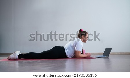 Young Plus Size Woman Stretching At Home Online. Flexible girl practices yoga and watches an online course on a laptop