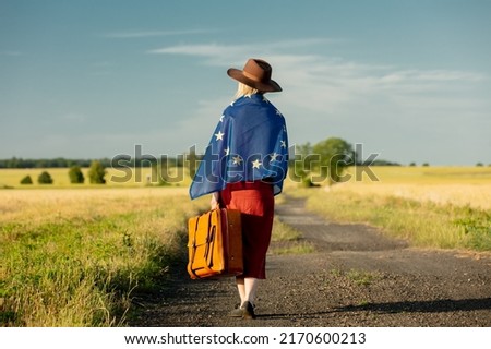 Girl in EU flag with suitcase on country road in sunset