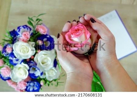 Female hands hold a rose flower. Notebook, diary with a pen on a wooden table near a bouquet of multi-colored roses in the background. Top view, flat lay.