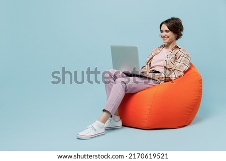 Full body young smiling happy woman 20s in brown shirt sit in bag chair hold use work on laptop pc computer isolated on pastel plain light blue background studio portrait. People lifestyle concept.